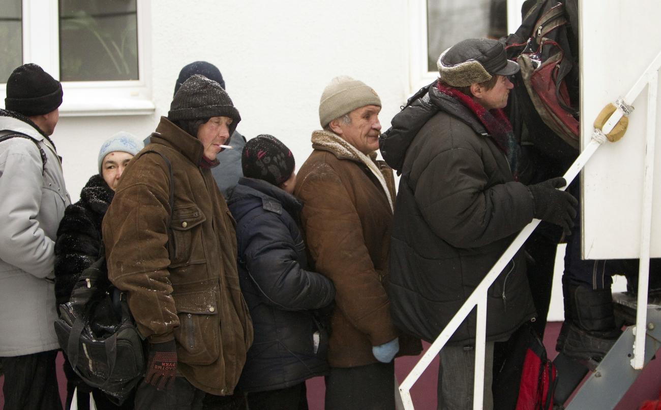 Picture shows a line of older men waiting in front of the steps of a clinic heavily bundled in winter wear against the cold.