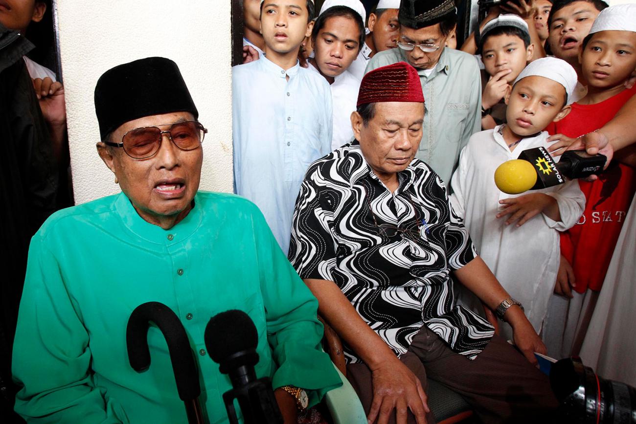 The sultan sits in a chair talking into microphones with the top of his cane resting against his chest. He is surrounded by followers, men and boys, all are wearing mostly white traditional Indonesian hats. The sultan’s hat is black, his shirt is a slate green, and he has amber-tinted lenses in his oversize glasses. He is leaning back and photographed in mid-sentence.