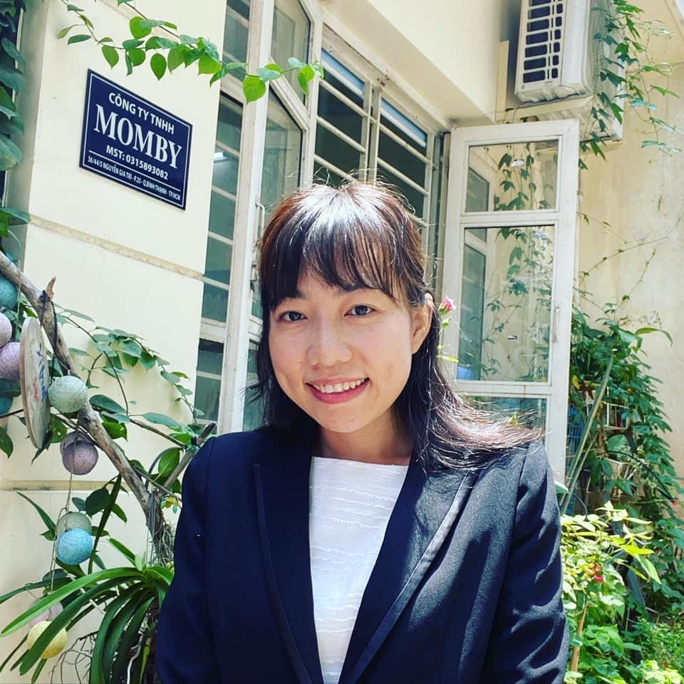 Ngoc Nguyen, a young woman in a black blazer stands in front of a yellow building with a French doors and a blue plaque that says Momby. The building is surrounded by lush green plants.