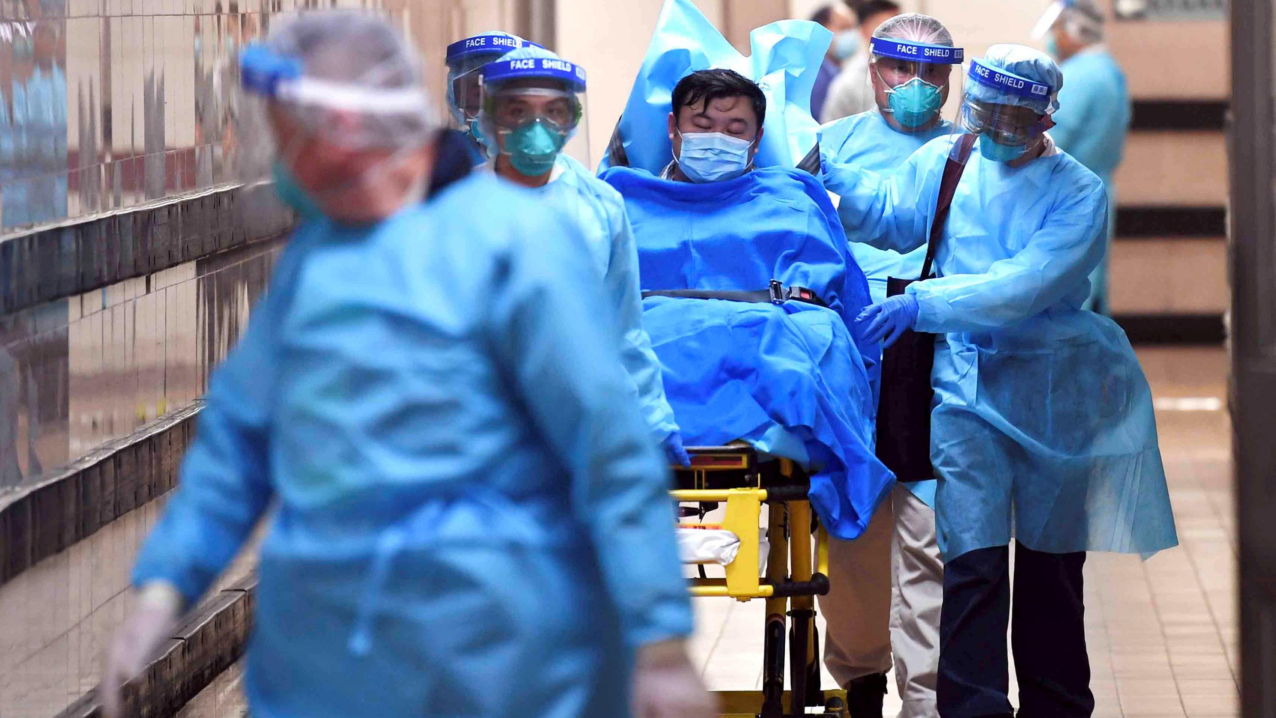 Picture shows a patient on a wheeled bed surrounded by four health care providers moving him with another caregiver walking in front. They all are covered head-to-toe in blue protective gear. 