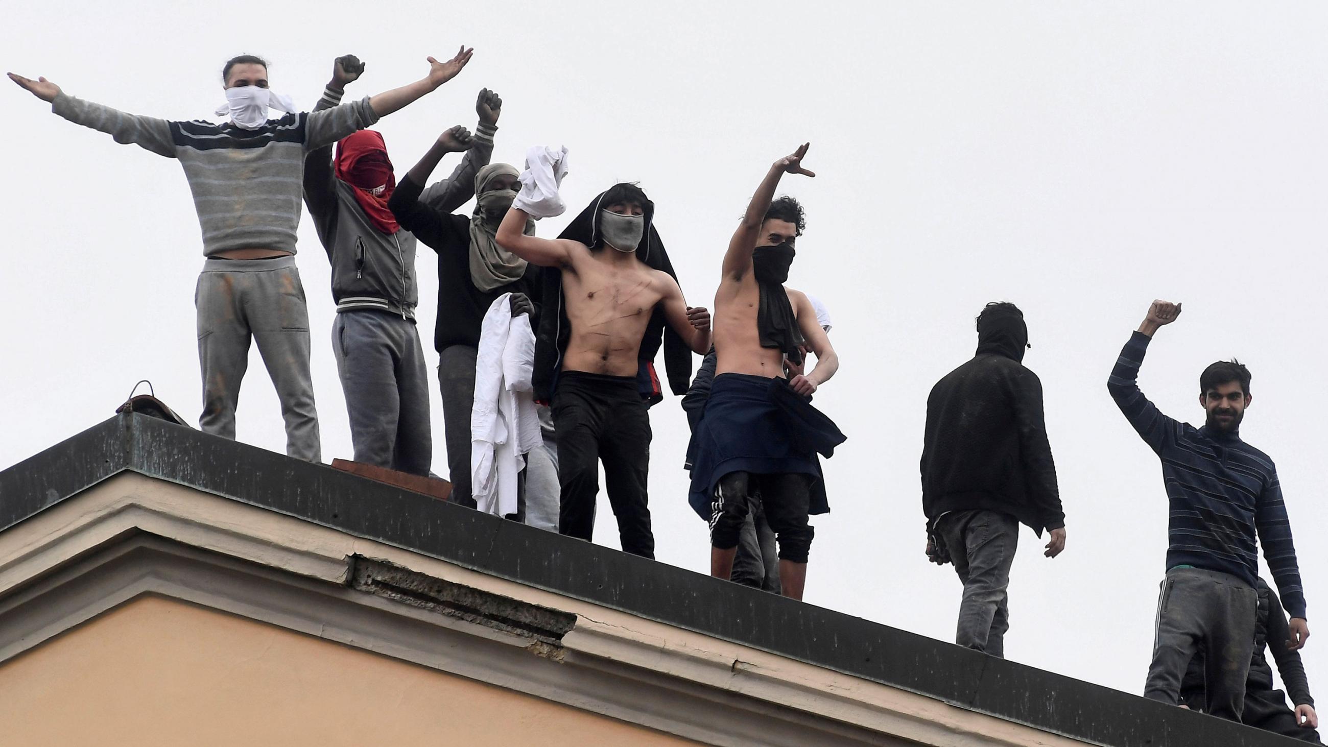 The photo shows a number of inmates, some with bandanas over their faces, on the roof of a prison building shouting at the camera, which appears to be some distance away. 