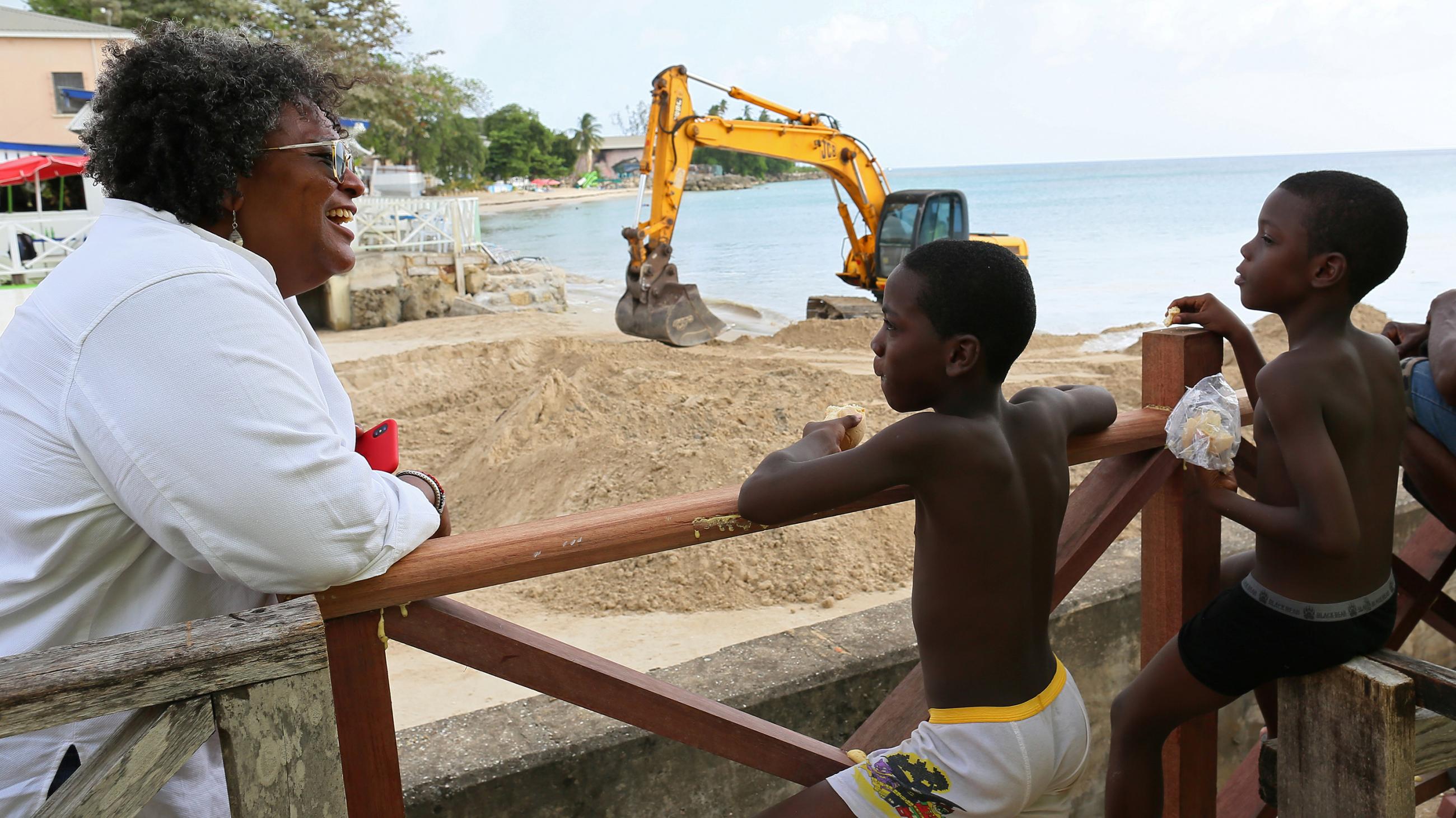 The photo shows the prime minister at the beach leaning on a rail talking to several young children. Construction machinery can be seen in the background. 