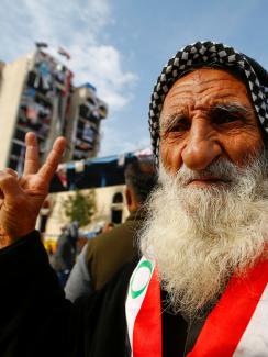 An elderly demonstrator poses for the camera as he makes a V-sign during ongoing anti-government protests, in Baghdad, Iraq, December 10, 2019. He has a black-and-white checkered scarf tied on his head and a red and white sash with green accents, around his neck. He has a very long white beard and friendly eyes. 