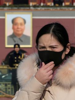 The photo shows a woman in a black mask in the foreground with a huge portrait of Chairman Mao visible in the background, along with a uniformed officer standing guard. 