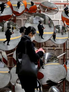 This is an amazing image of a woman walking by a display of spherical mirrors, the sort of which someone might place on a corner to see a wide angle. She is wearing a black face mask and is reflected in all the mirrors.