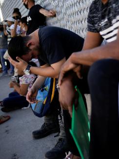 This is a powerful image showing people lined up seated with their backs to a chain-link fence covered in barbed wire. Some of them hold packets of papers. One of them has his head resting in his hands.