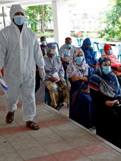 The photo shows a crowd of people wearing masks waiting on line in chairs while a health worker in full protective gear walks beside them. 