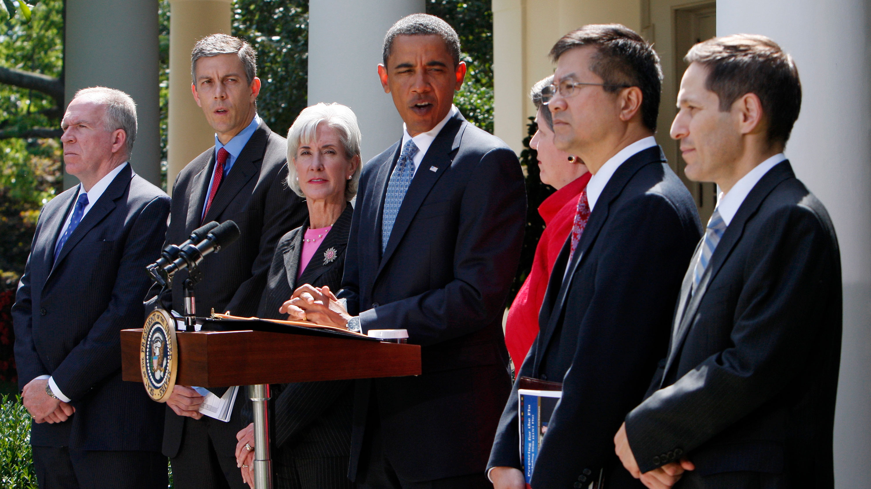 Obama is standing in front of microphones looking toward the camera surrounded by cabinet officials. 