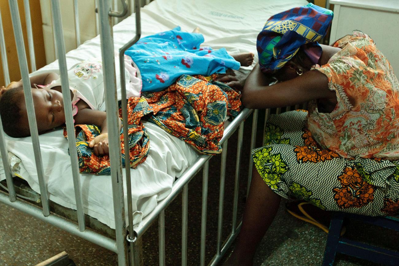 Image shows a young child in a hospital crib sleeping on her back with her arms op splayed on each side of her head. A woman, presumably her mother or caregiver, sits beside the bed with her head down, resting in her folded arms as she leans forward resting her head on the bed. Is her head down from exhaustion or despair? The photograph cannot answer that question.