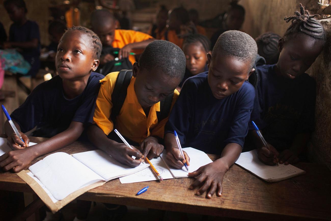 The picture shows a crowded classroom lit with natural light and several students crammed at a wooden desk with heads down and pen to paper. One student, at the end of the desk can be seen looking up, perhaps pondering what to write next.