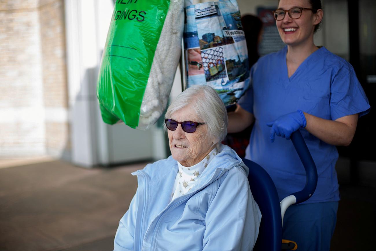 The photo shows the nurse wheeling the patient who is wearing dark sunglasses. The nurse is smiling broadly. 