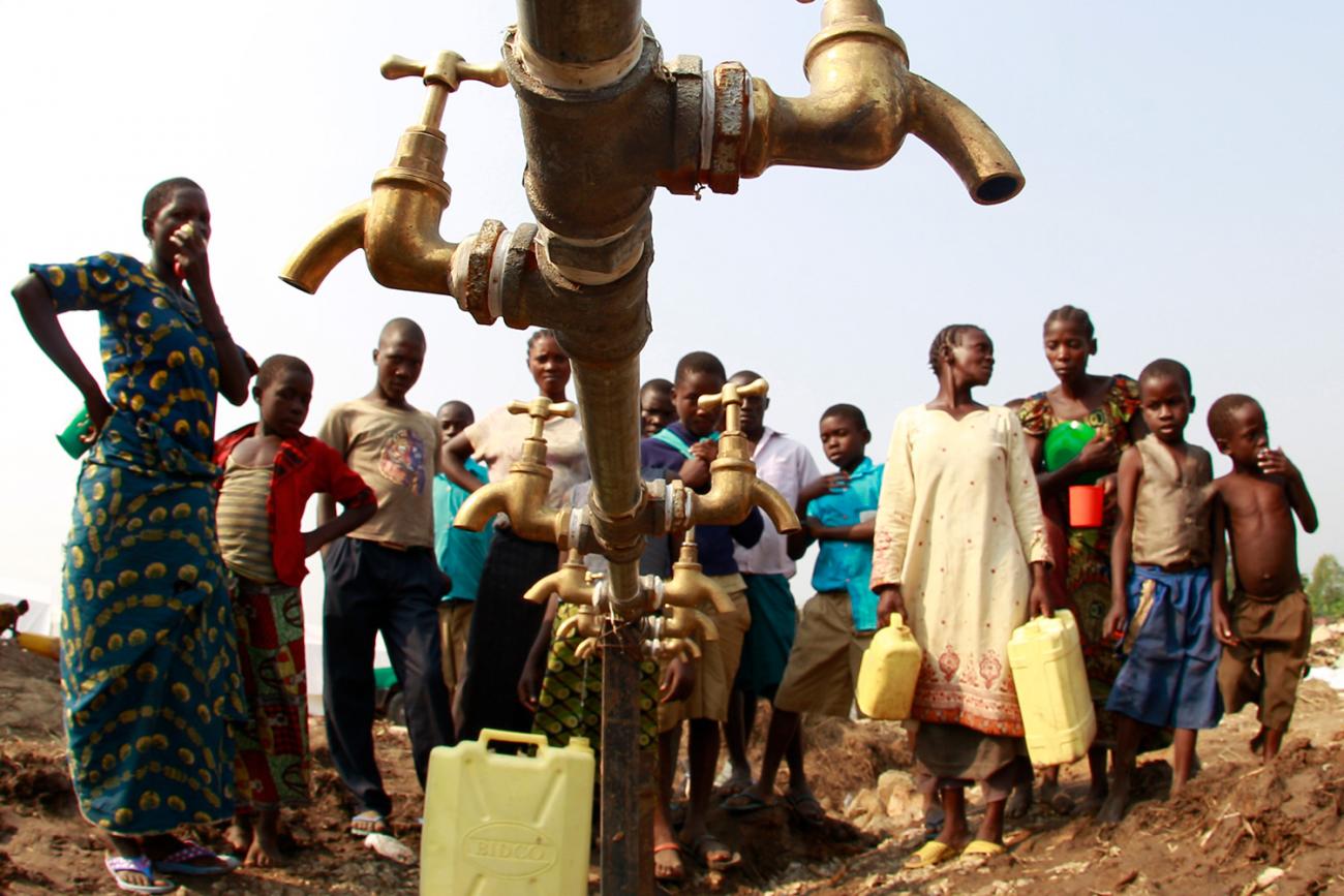 The photo shows a crowd of people around a metal water pipe with no water flowing. 