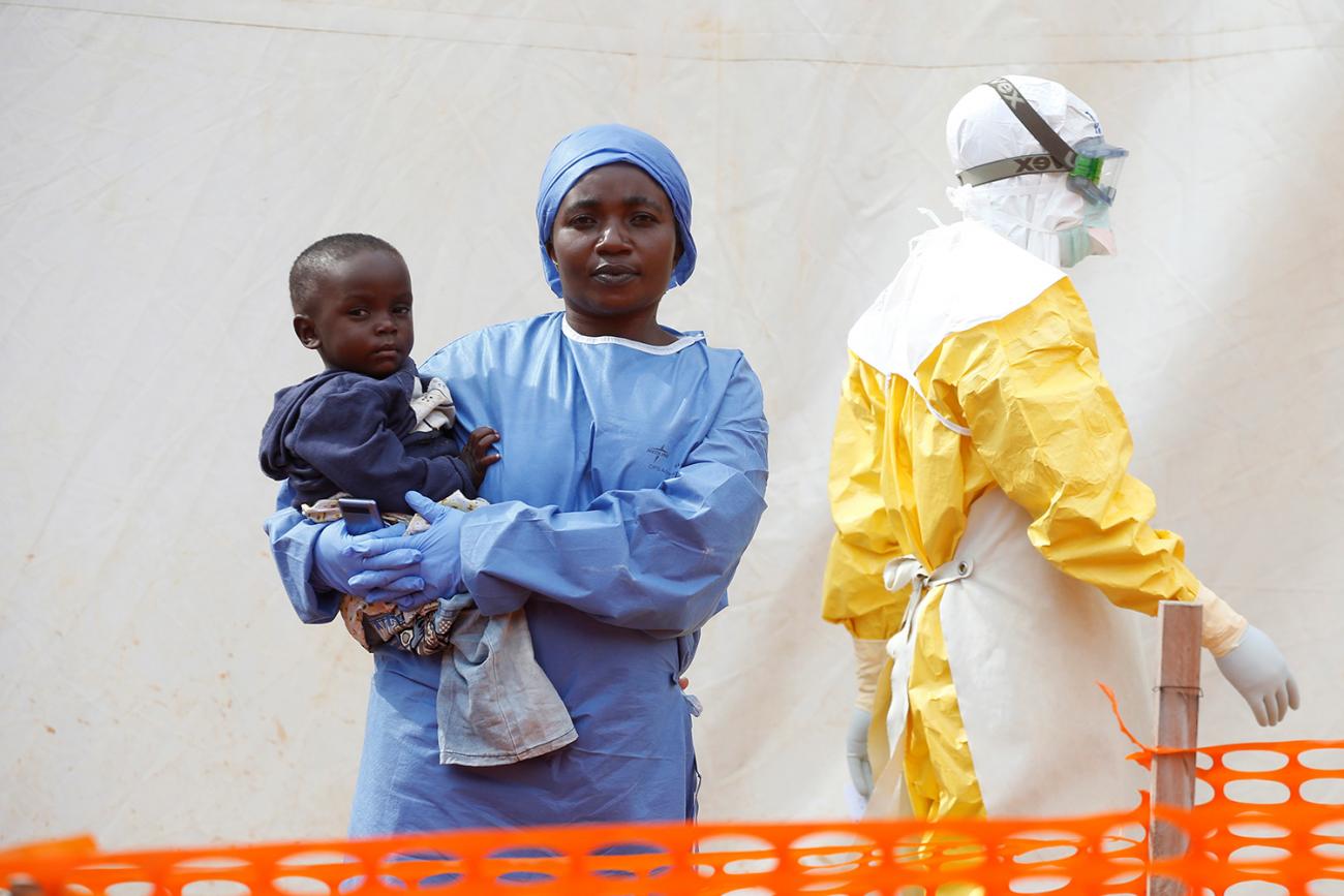 Picture shows a woman dressed in blue hospital scrubs holding a baby. A man in a full protective yellow suit is walking by in the background. 