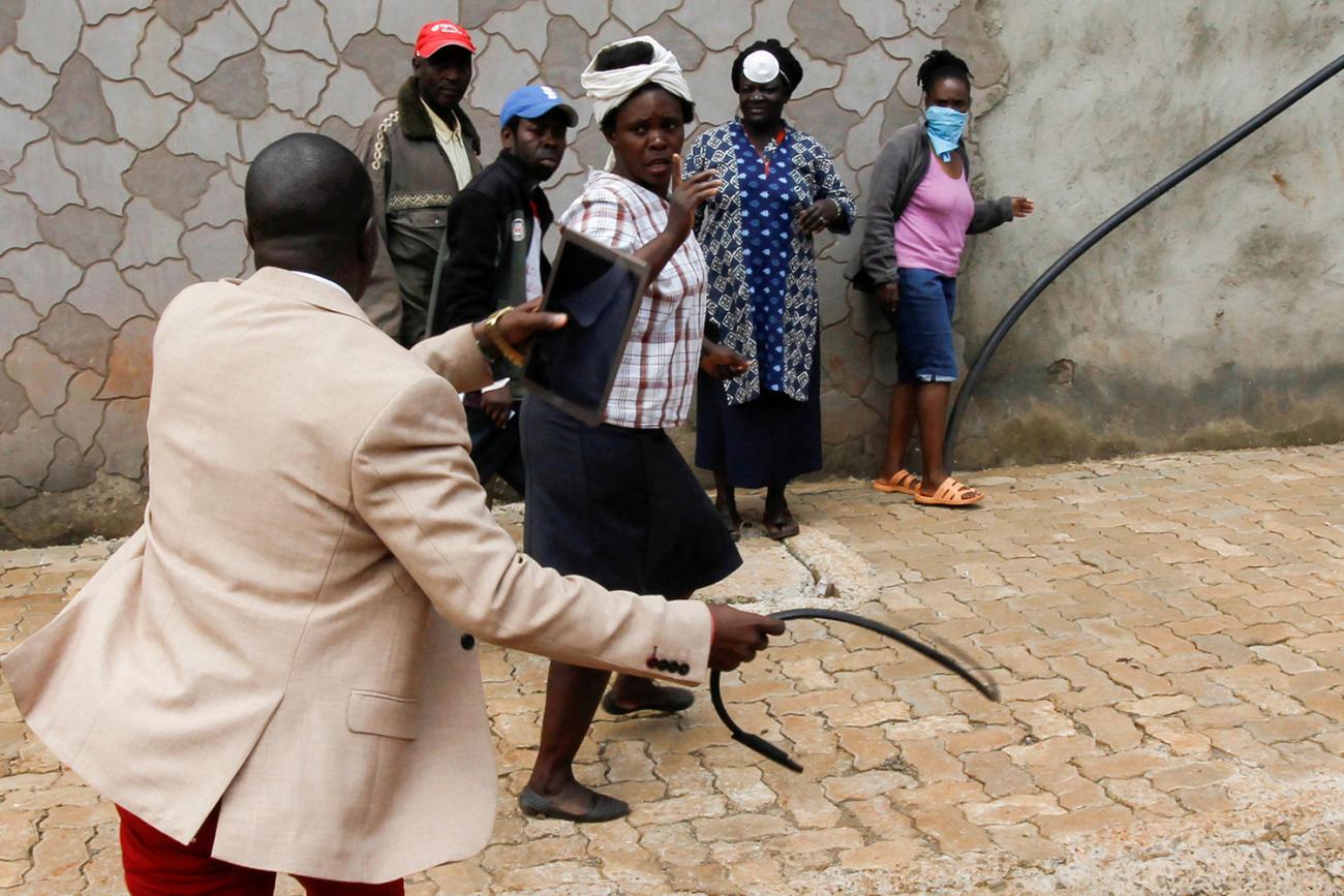 The photo shows a man in a jacket with his back to the camera in motion burnishing a black whip of some sort in his right hand while three women and two men who are walking away, apparently under threat. 