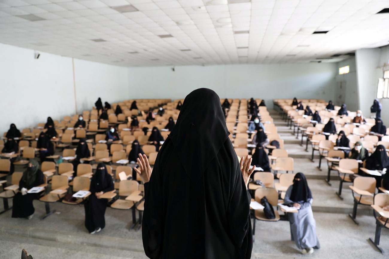 Picture shows a large classroom-like space filled with women who are practicing social distancing by sitting in every other row an every other desk. One woman in front is lecturing. 