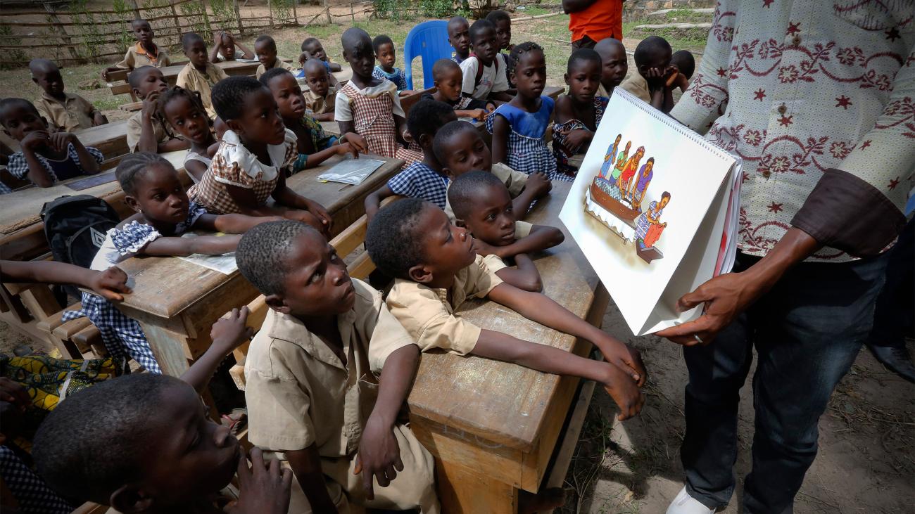 The photo shows the teacher at the front of a room showing a placard to the children. 