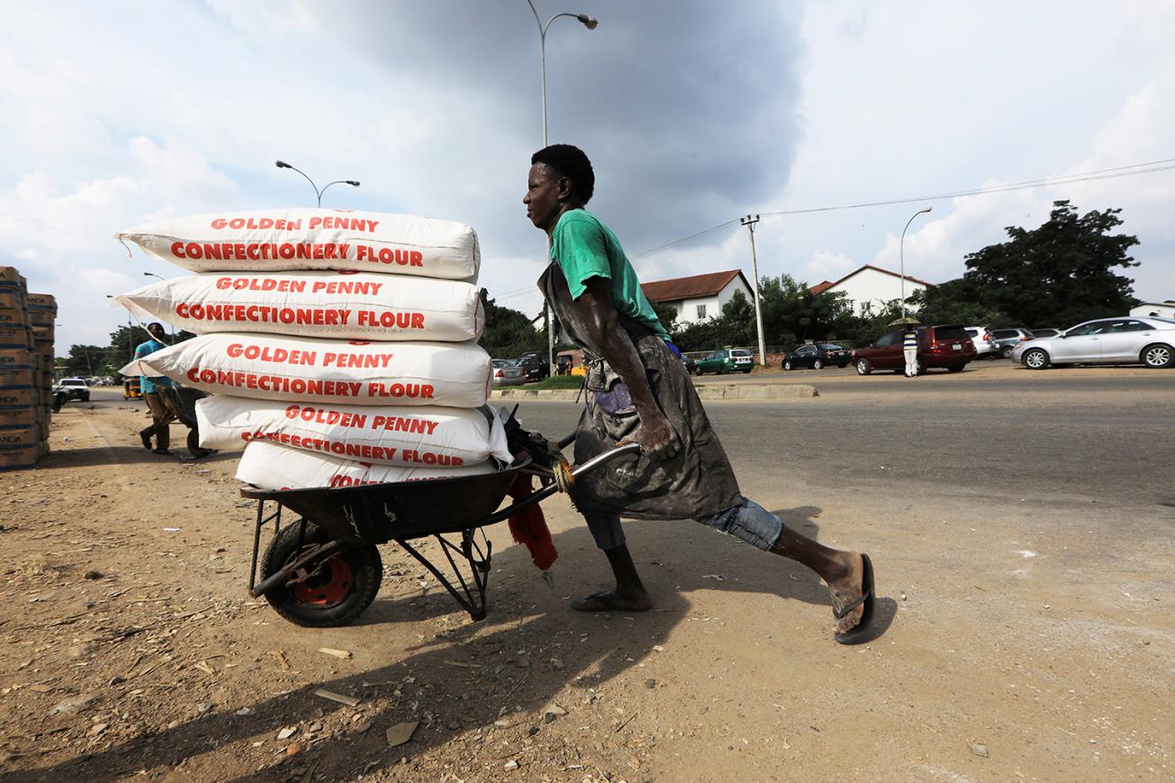 The photo shows a man wheeling a barrow stacked high with what appear to be very heavy bags, probably hundreds of pounds total. 