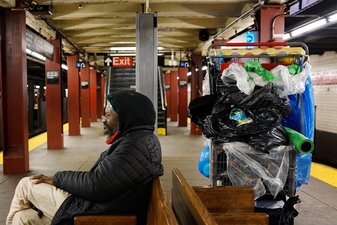 Picture shows a man sitting on a bench with large cart of stuff sitting behind him. 