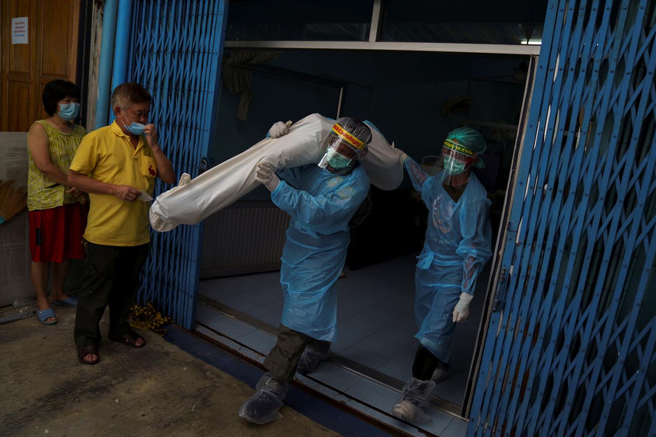 The photo shows two workers in blue protective gear walking out of a building. One of them has a body wrapped in a white covering over his shoulder. 