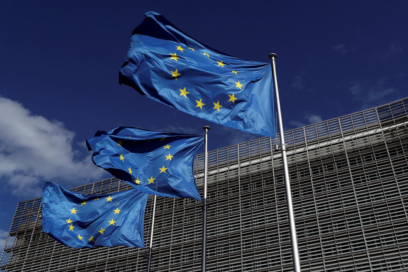 The photo shows the glass and steel building against a deep blue sky with European Union flags in the foreground. 