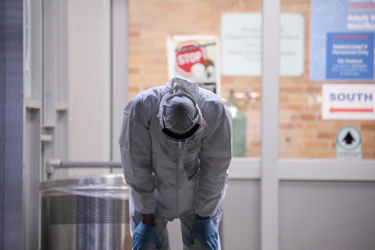 Picture Shows a medical worker standing in full protective gear outside a hospital, bending over. 