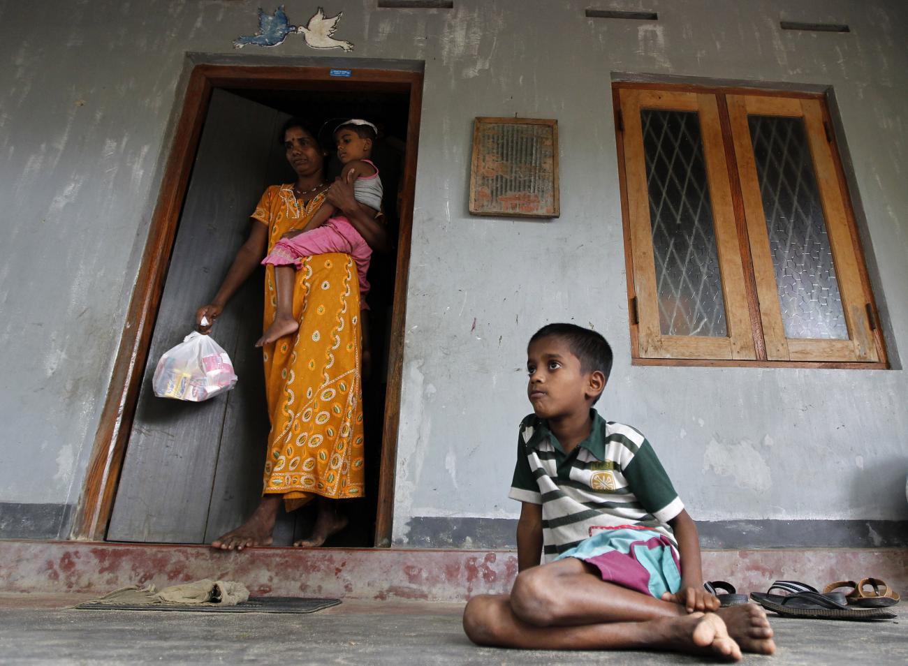 Mother holding child comes out a door, while a young boy sits on the floor. 