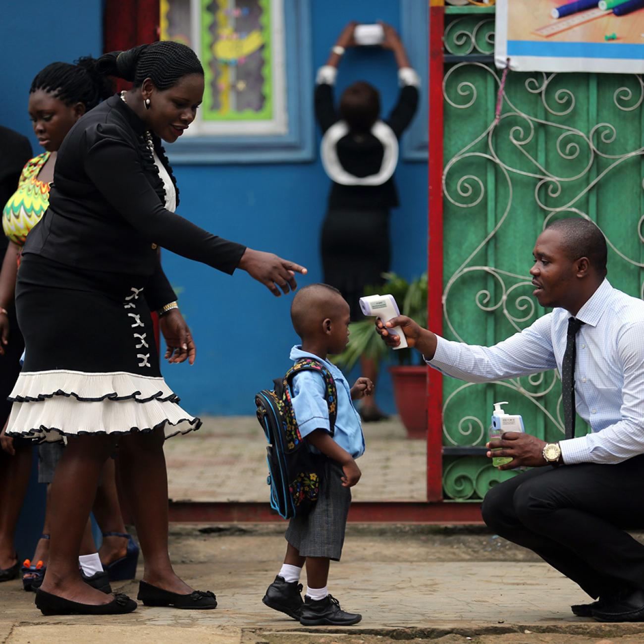 Picture shows a young boy carrying a backpack and wearing a school uniform standing in front of a brightly-colored building. A well-dressed woman directs him while a well-dressed man kneels and points a high-tech thermometer at his head to take his temperature.