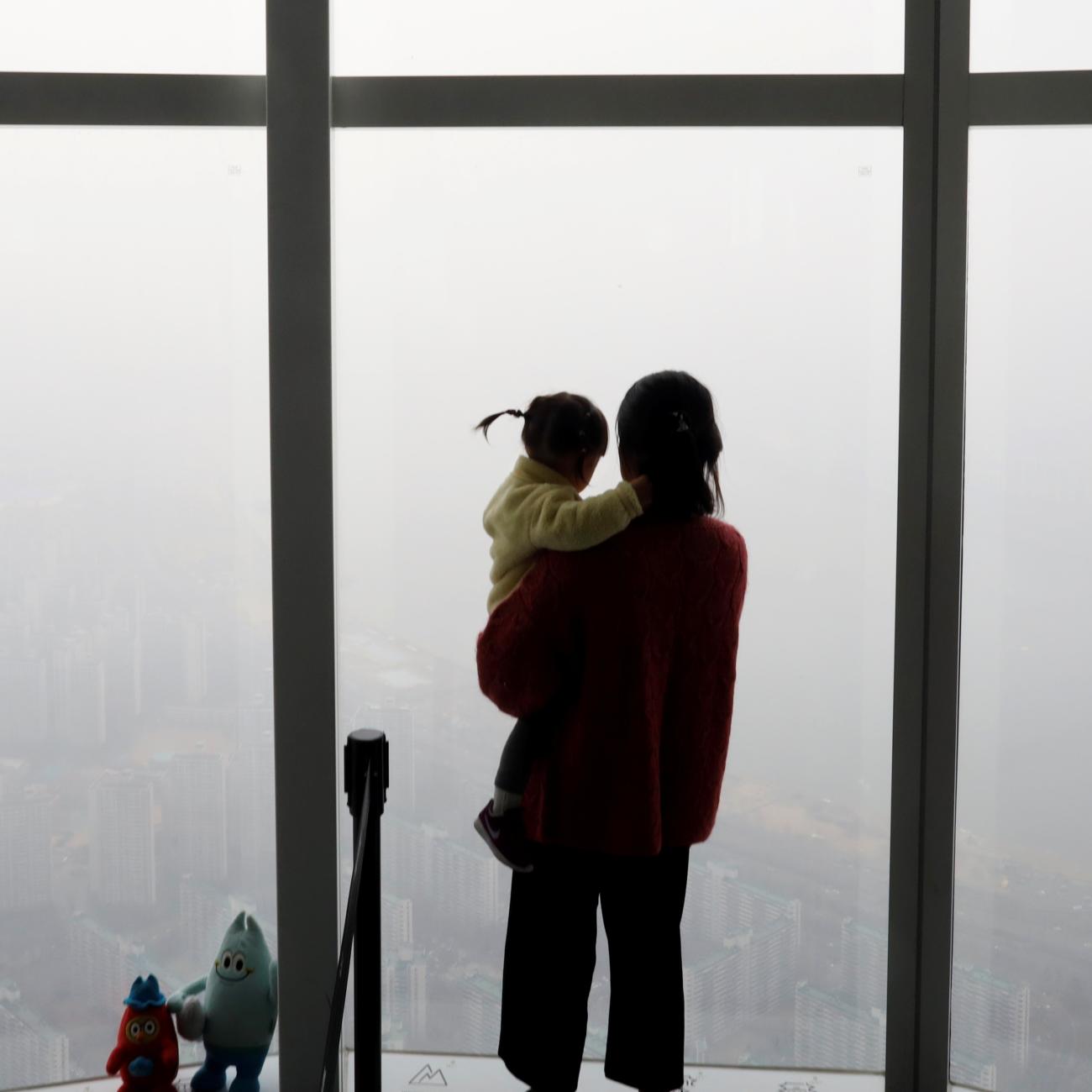 A woman holding her baby in her arms looks out a window at the city shrouded by a fine dust during a polluted day.