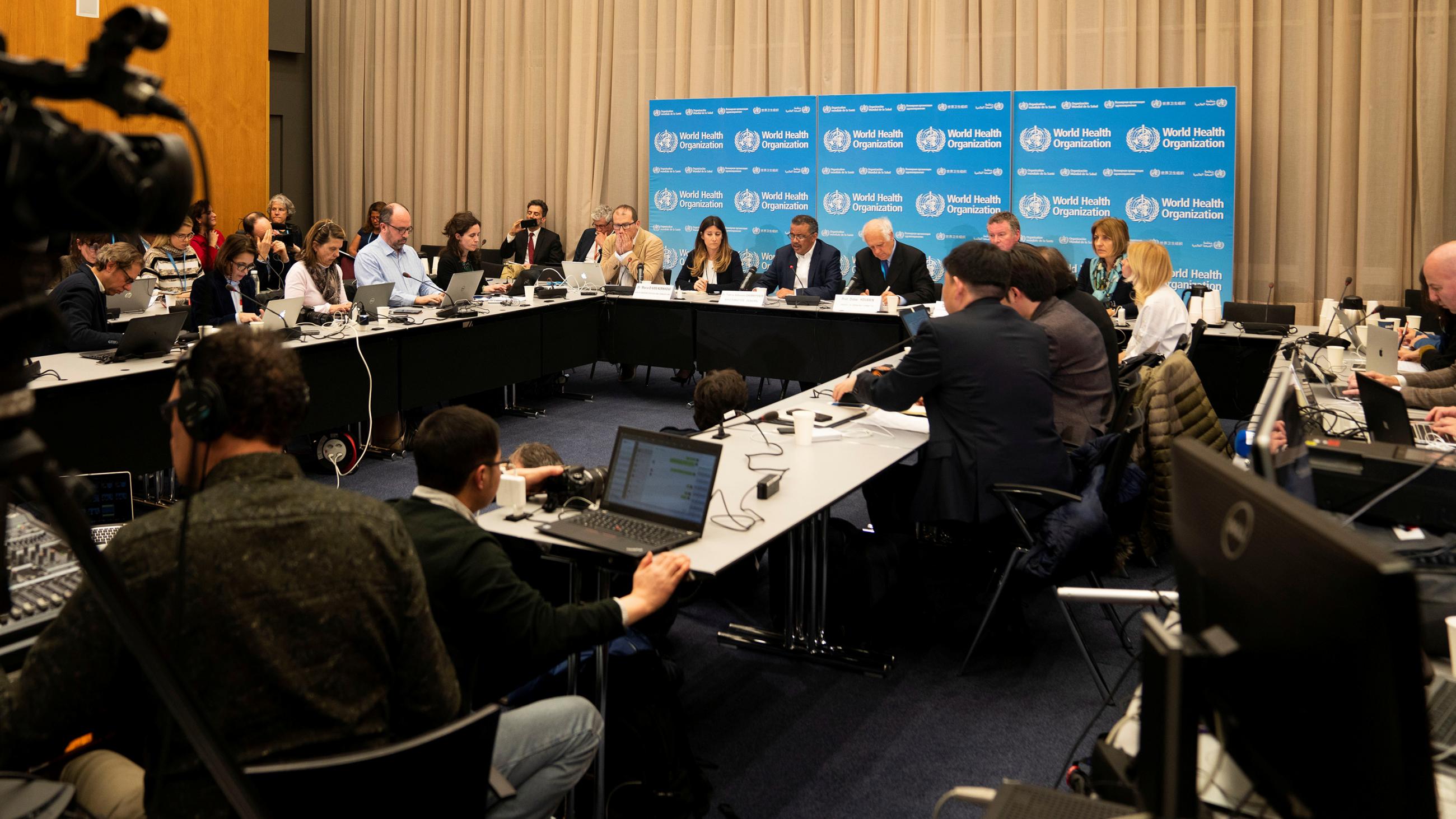 Picture shows a crowded press conference with officials seated at a long table and lots of reporters in the room. 