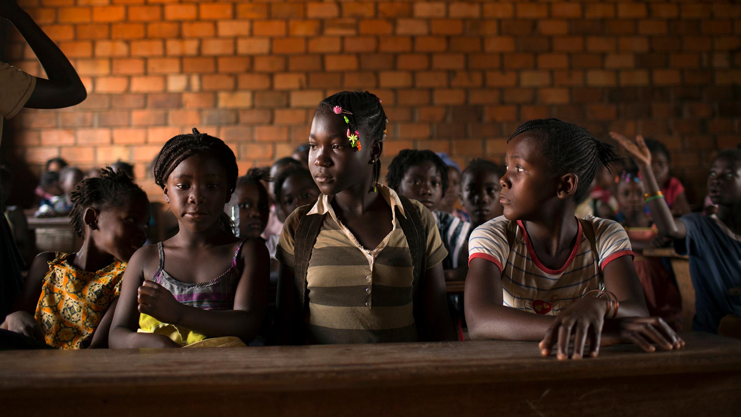 Photo shows a classroom packed with pre-teen girls sitting at desks in front of a brick wall. 