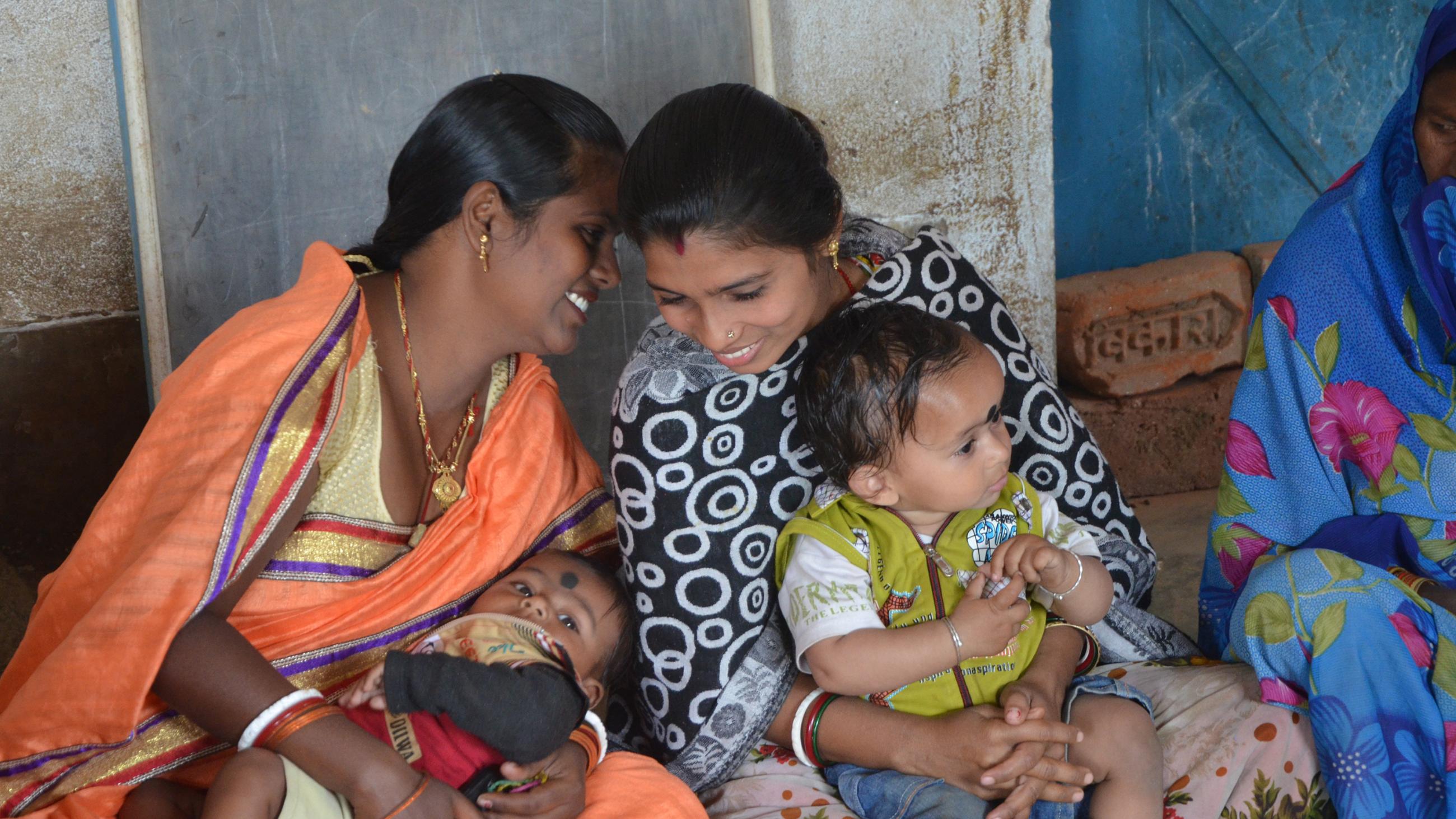 The photo shows two women seated holding small children on their laps smiling broadly as the talk. 