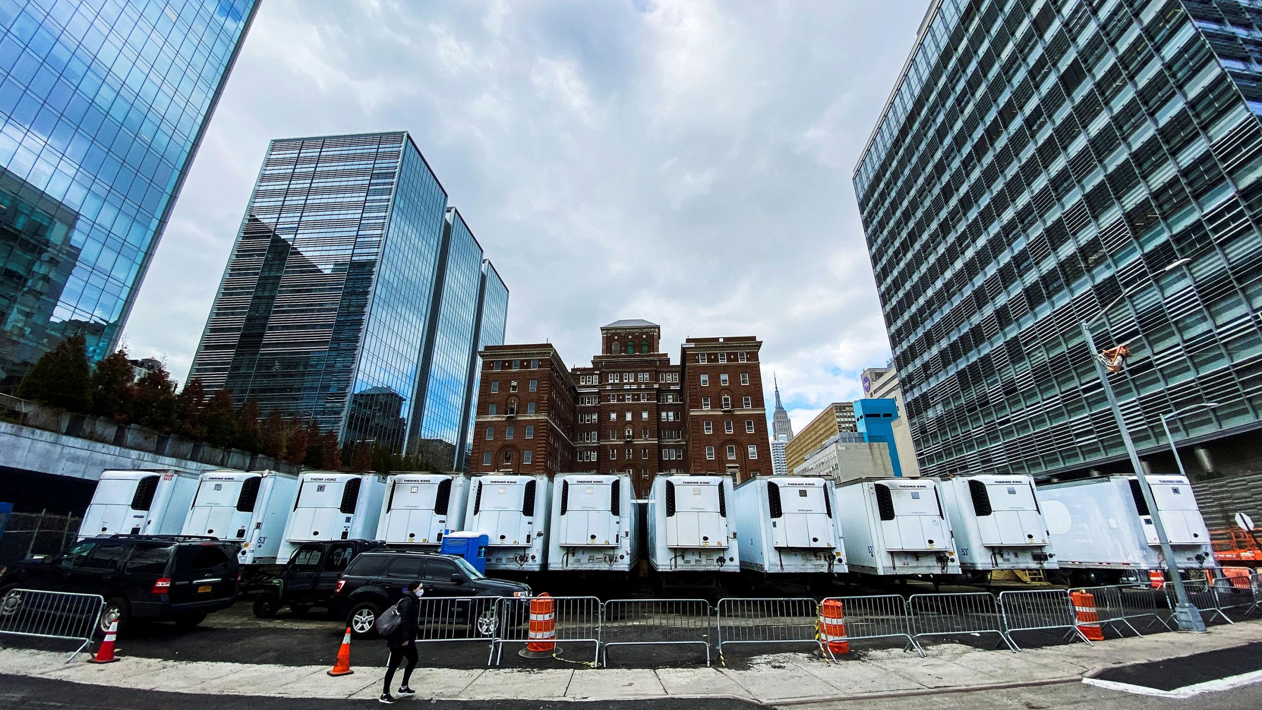 The photo shows a large hospital complex with a large line of a dozen refrigerated trucks. 