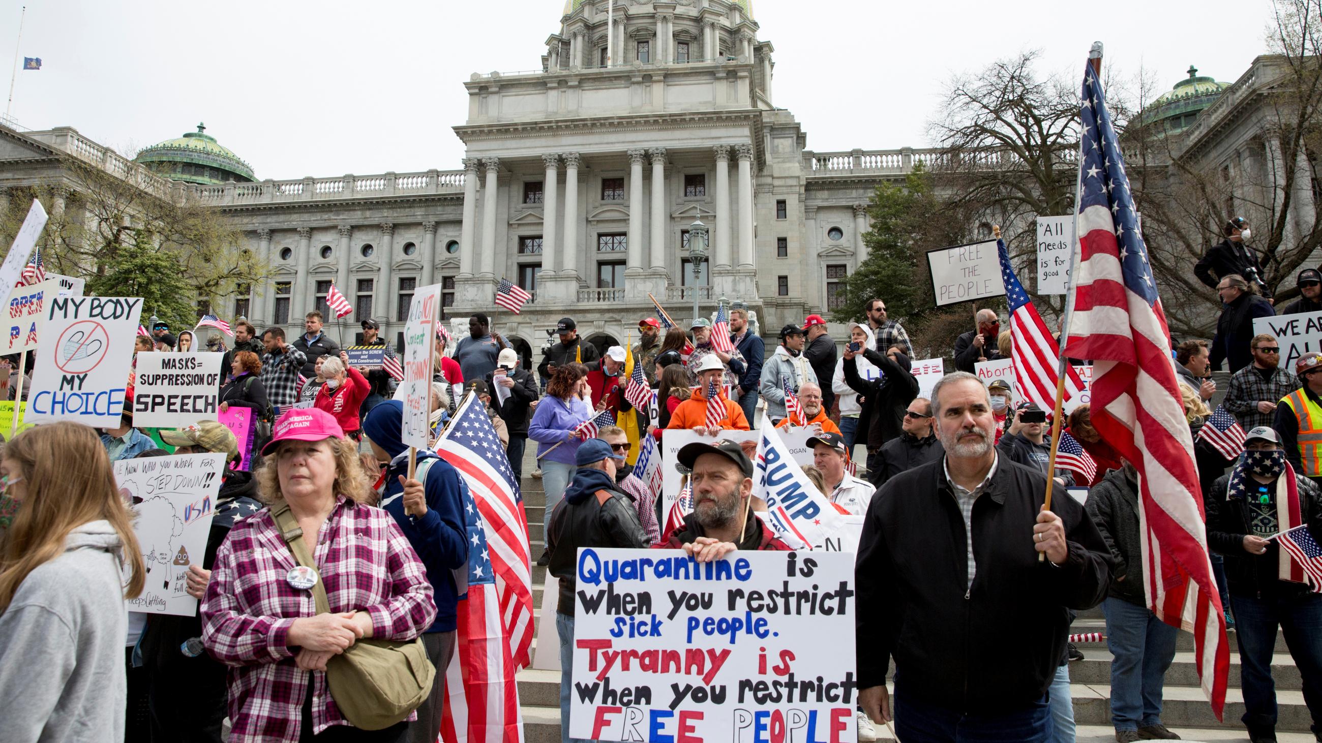  The photo shows a large number of protestors holding signs and waving the American flag on the steps of what appears to be the PA state house. Very few of them are wearing masks, and many of them are not practicing physical distancing. 