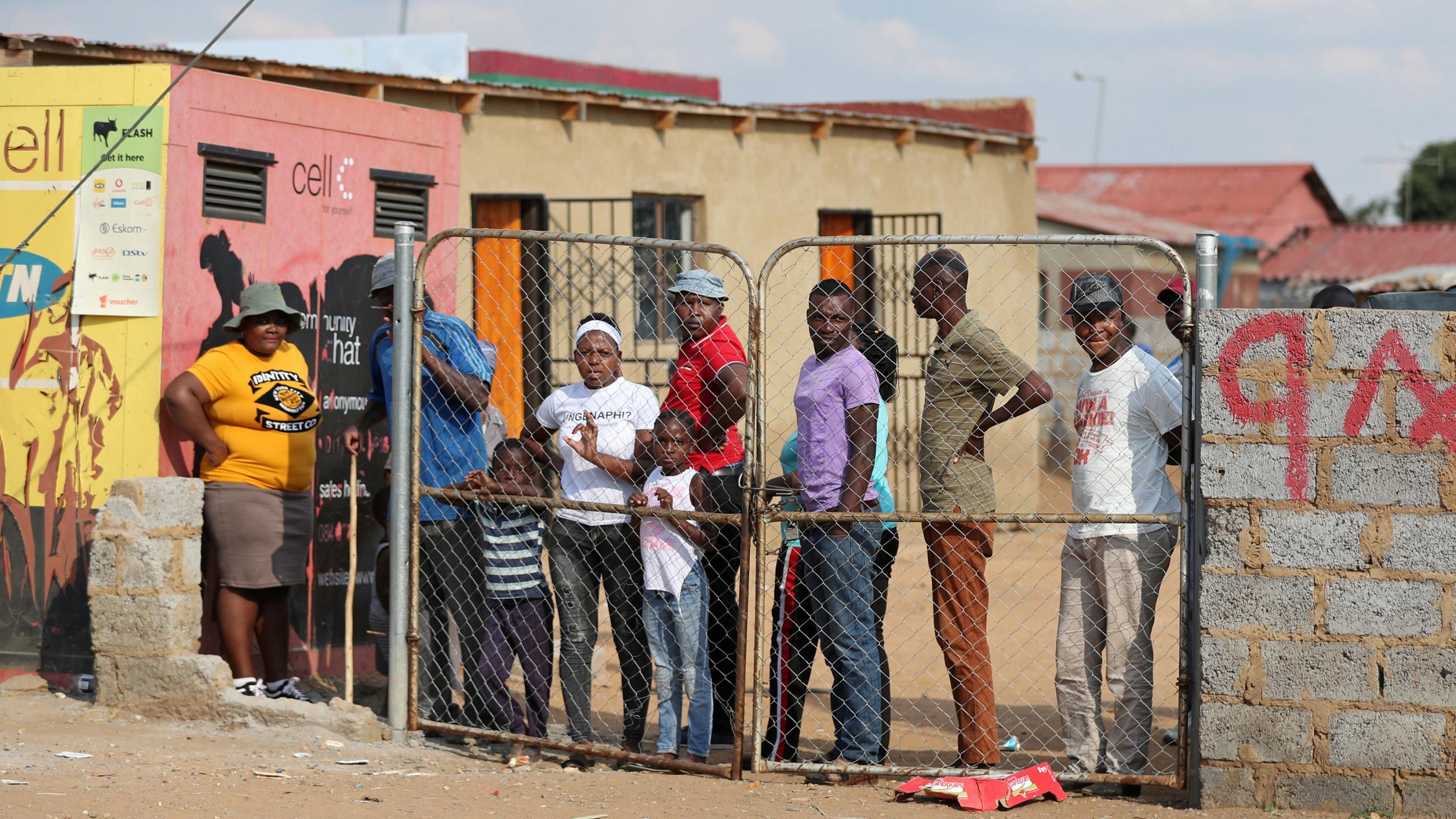 The photo shows several people congregated behind a closed fence though there is a very large gap between the fence and the adjacent wall. 