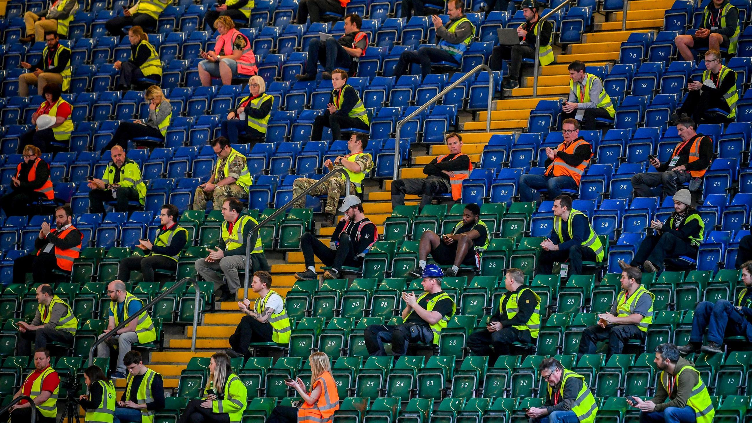 Picture shows emergency responders wearing yellow vests and sitting amid seats of a stadium. 