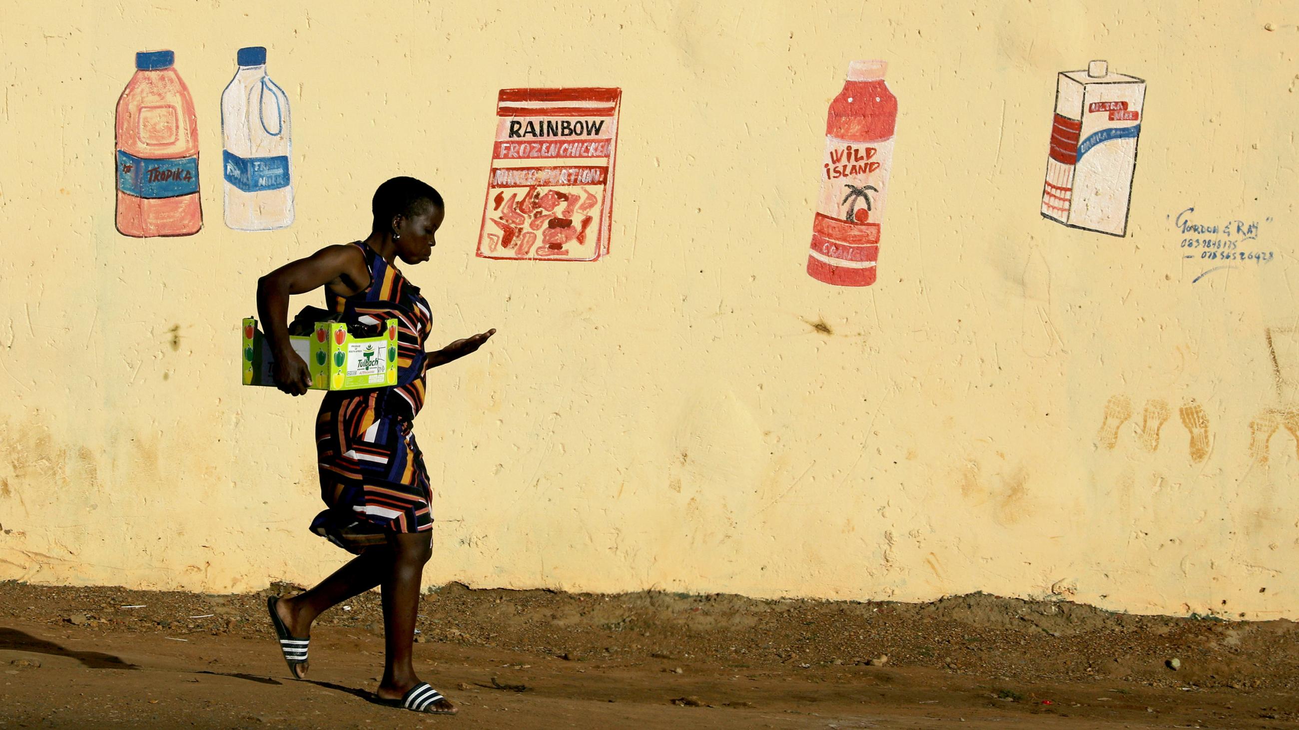  This is a striking photo of a woman carrying a box tucked under one arm and counting coins in the other walking in front of a butter yellow wall with faded, hand-drawn grocery items. 