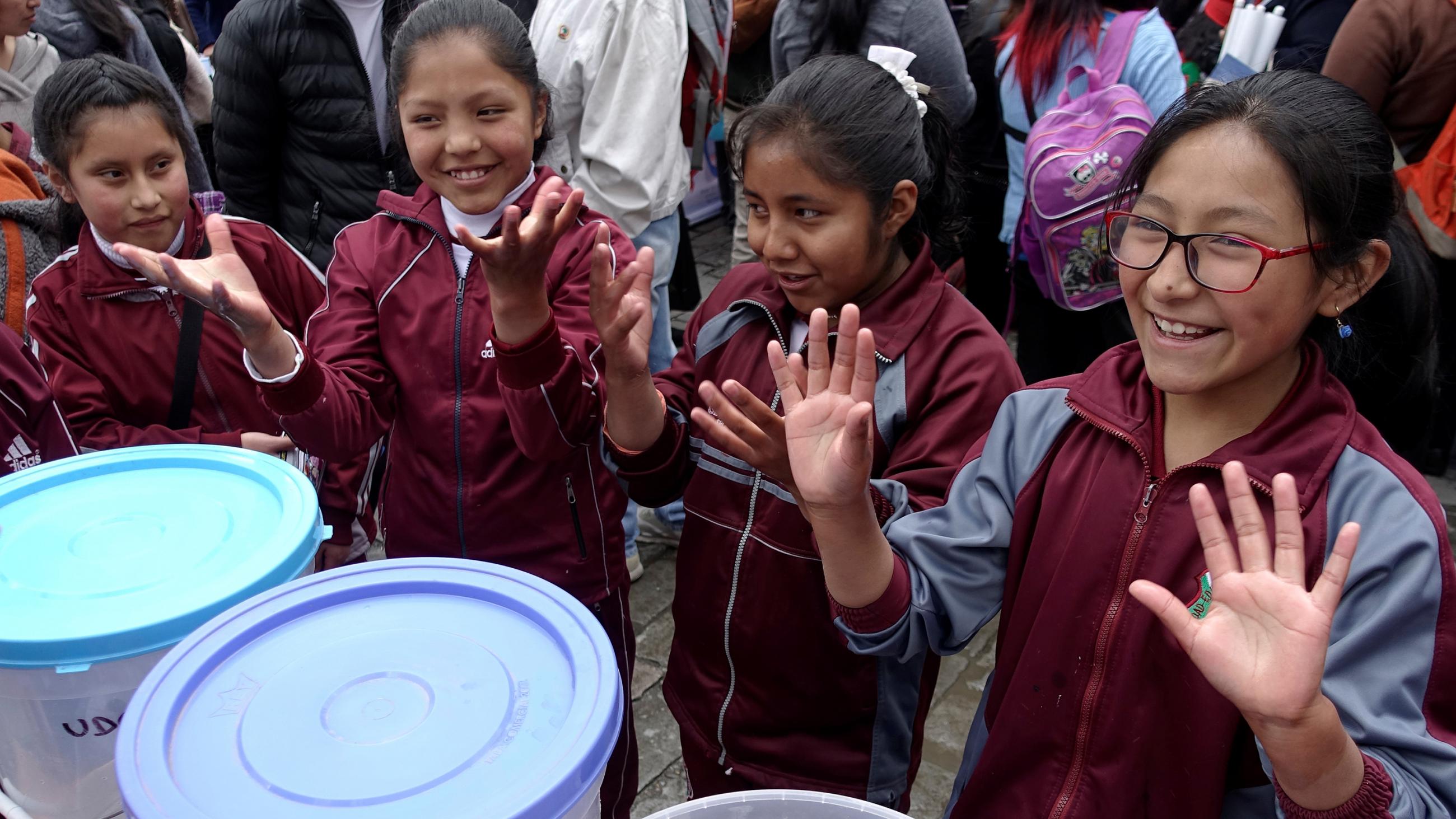 The photo shows a crowd of kids and adults. Four girls in school uniforms line up and show their hands to the camera. 