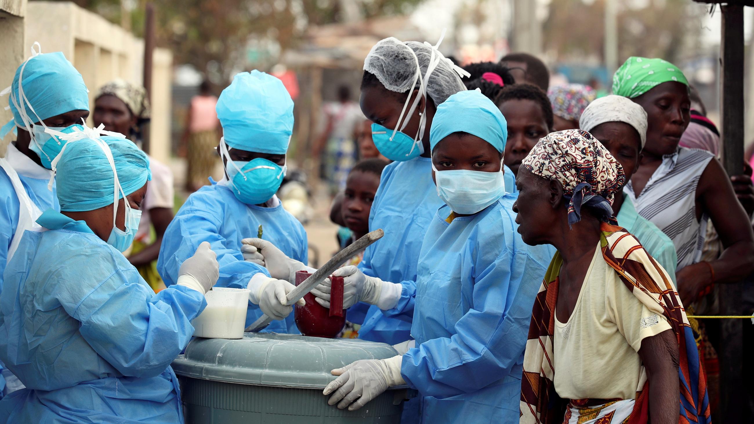 The photo shows a number of people wearing blue protective gear working on mixing a solution with a number of others wearing plain clothes looking on. 