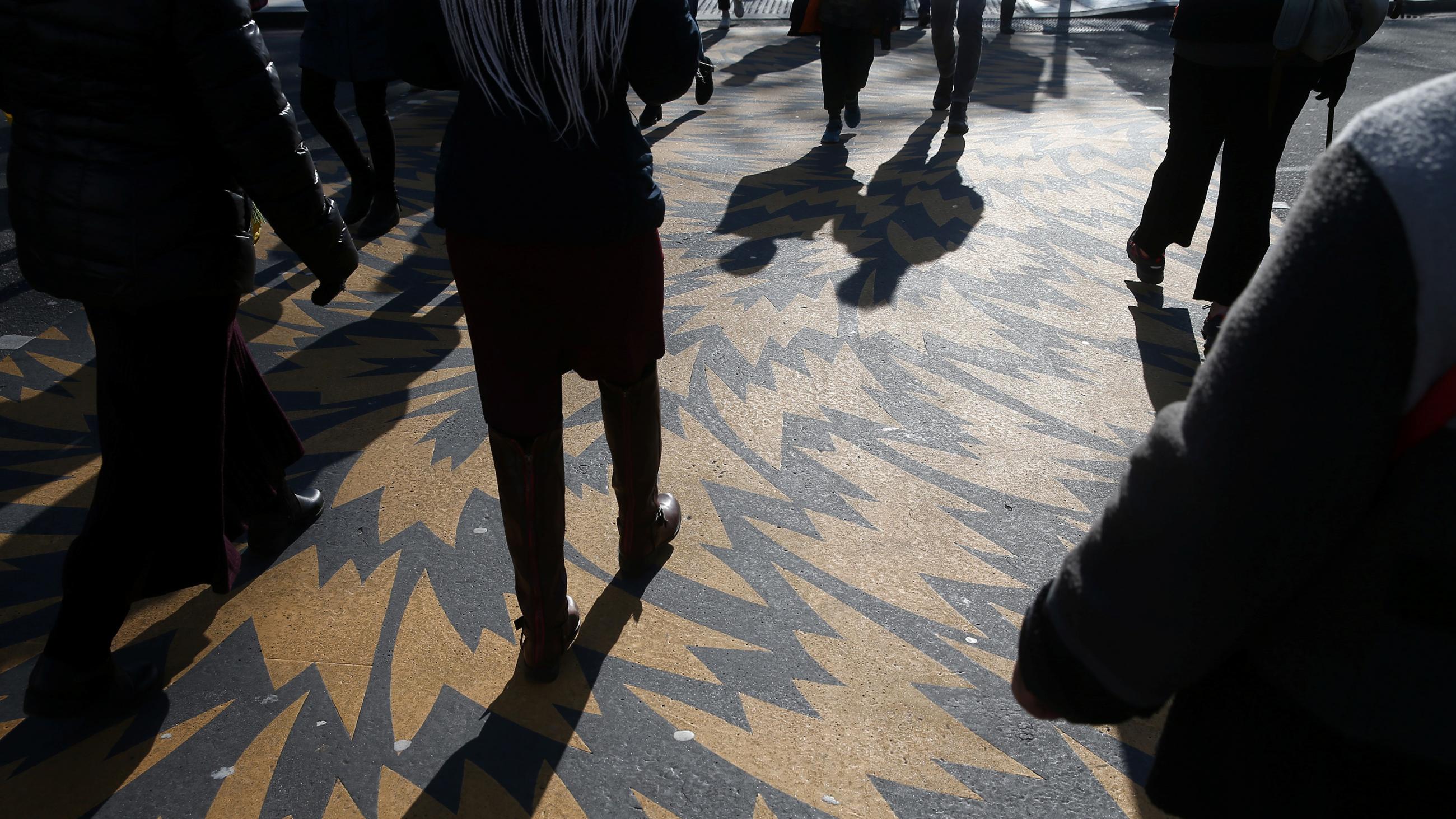 The photo shows the torso and legs of several people walking across a street over a decorative swirl pattern of paint on the crosswalk. 