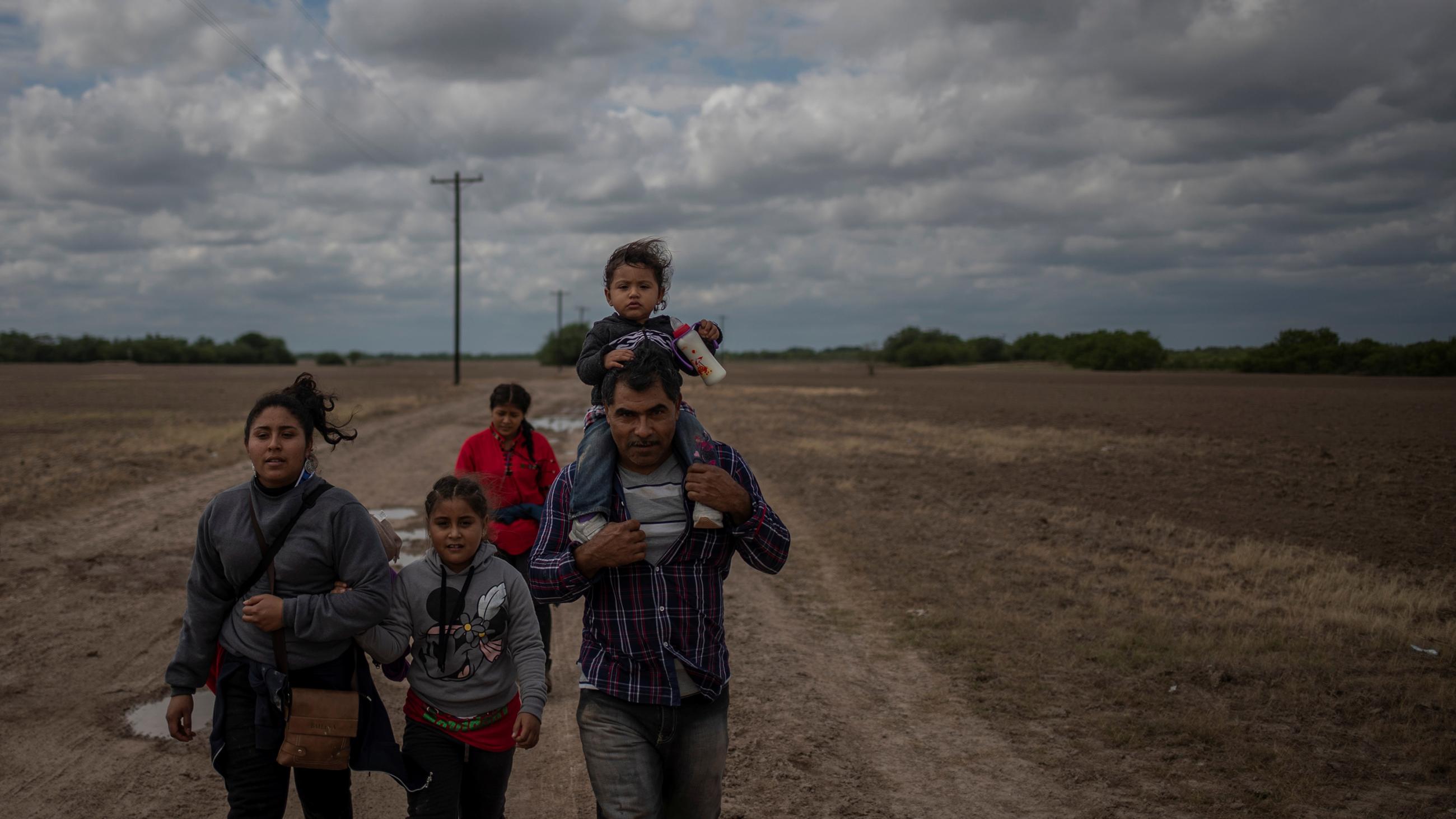 The photo shows a family walking through a field at dusk. The father has a small child on his shoulders. 