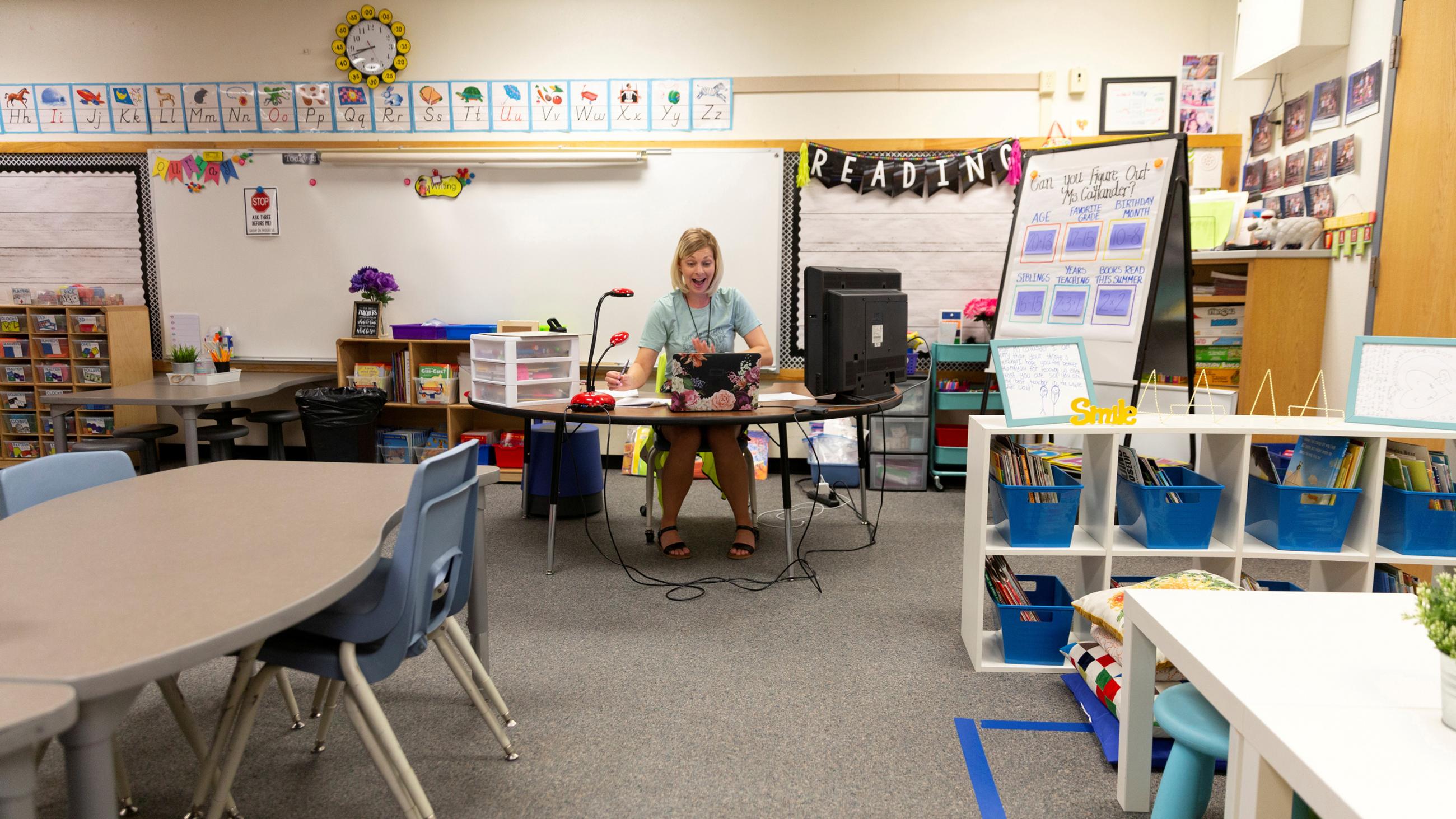 The photo shows the teacher in an empty classroom speaking expressively into the computer. 