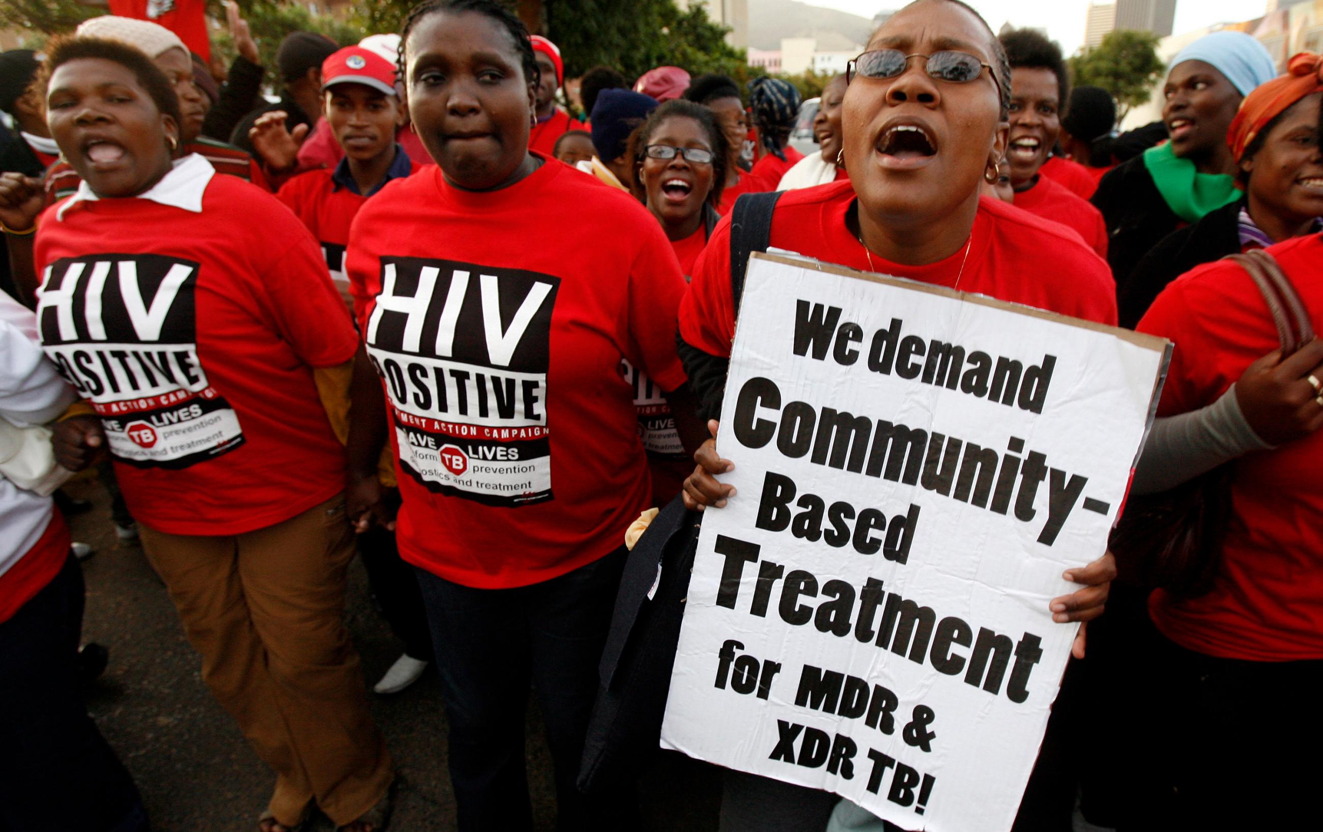 The photo shows a crowd of people wearing protest shirts and carrying signs calling for new community-based treatments. 