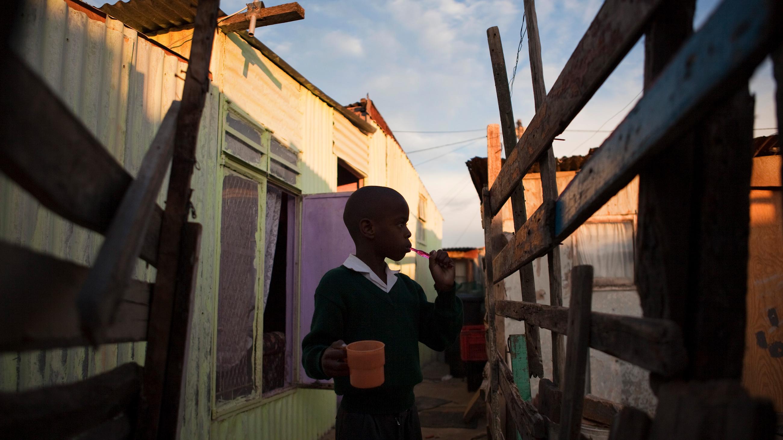 The photo shows Sinelizwi in an outdoor setting surrounded by ramshackle wooden structures. 