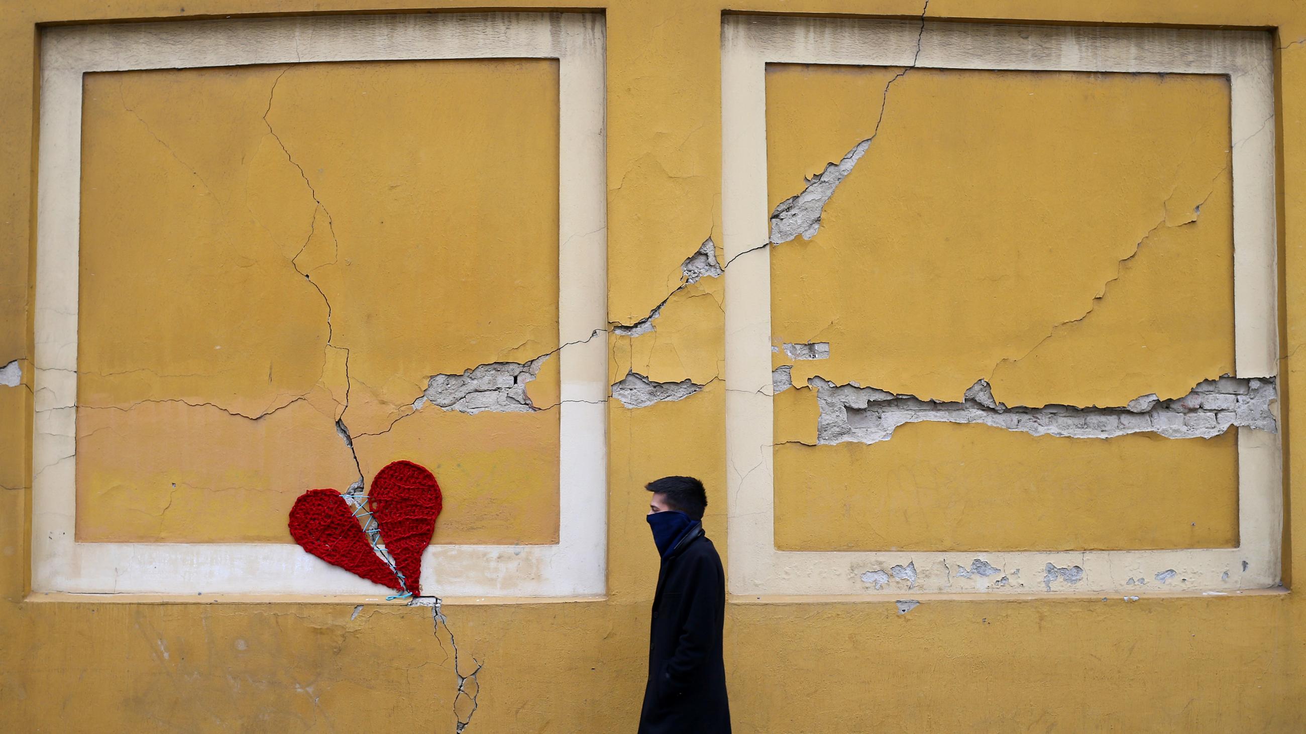 The photo shows a man walking past the heart on the side of a building that appears cracked. 