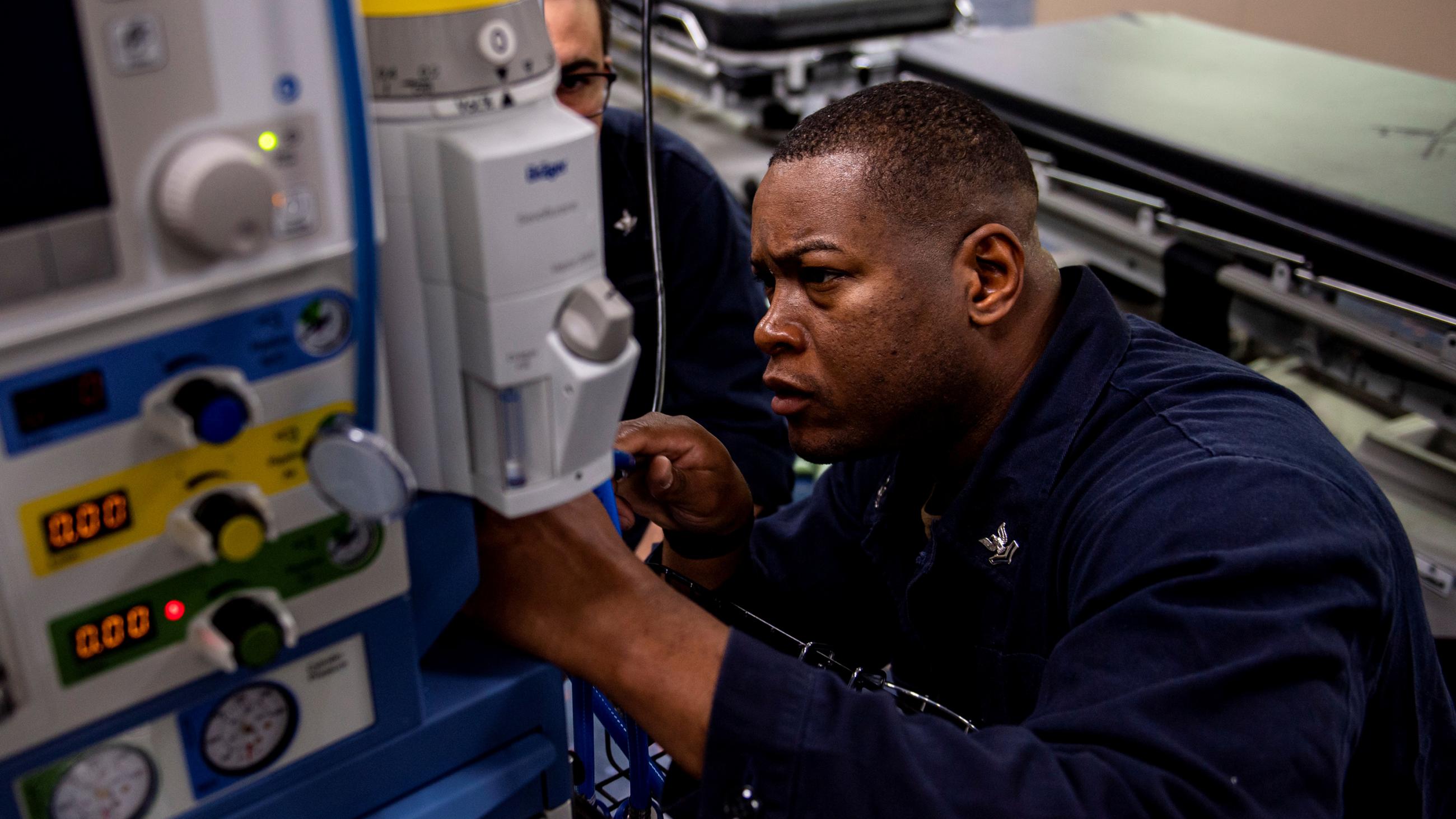 Picture shows the corpsman kneeling slightly while he works on a machine. 