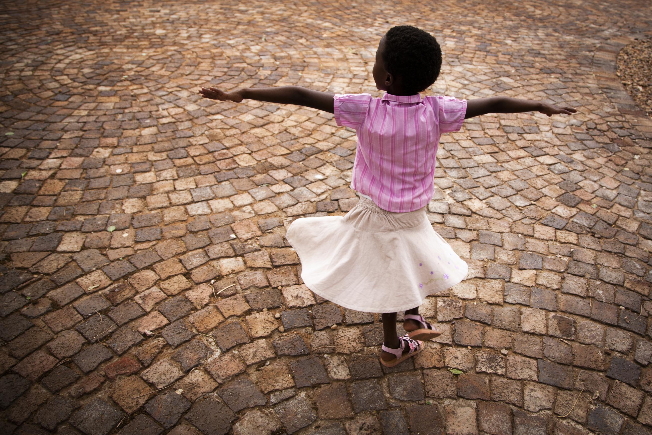 A little with her back turned to us is dressed in a pink shirt and white skirt. She twirls around on a brick-coverered street.