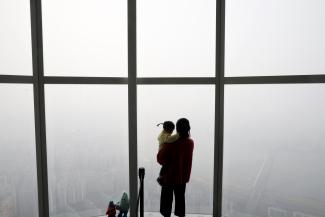 A woman holding her baby in her arms looks out a window at the city shrouded by a fine dust during a polluted day. 